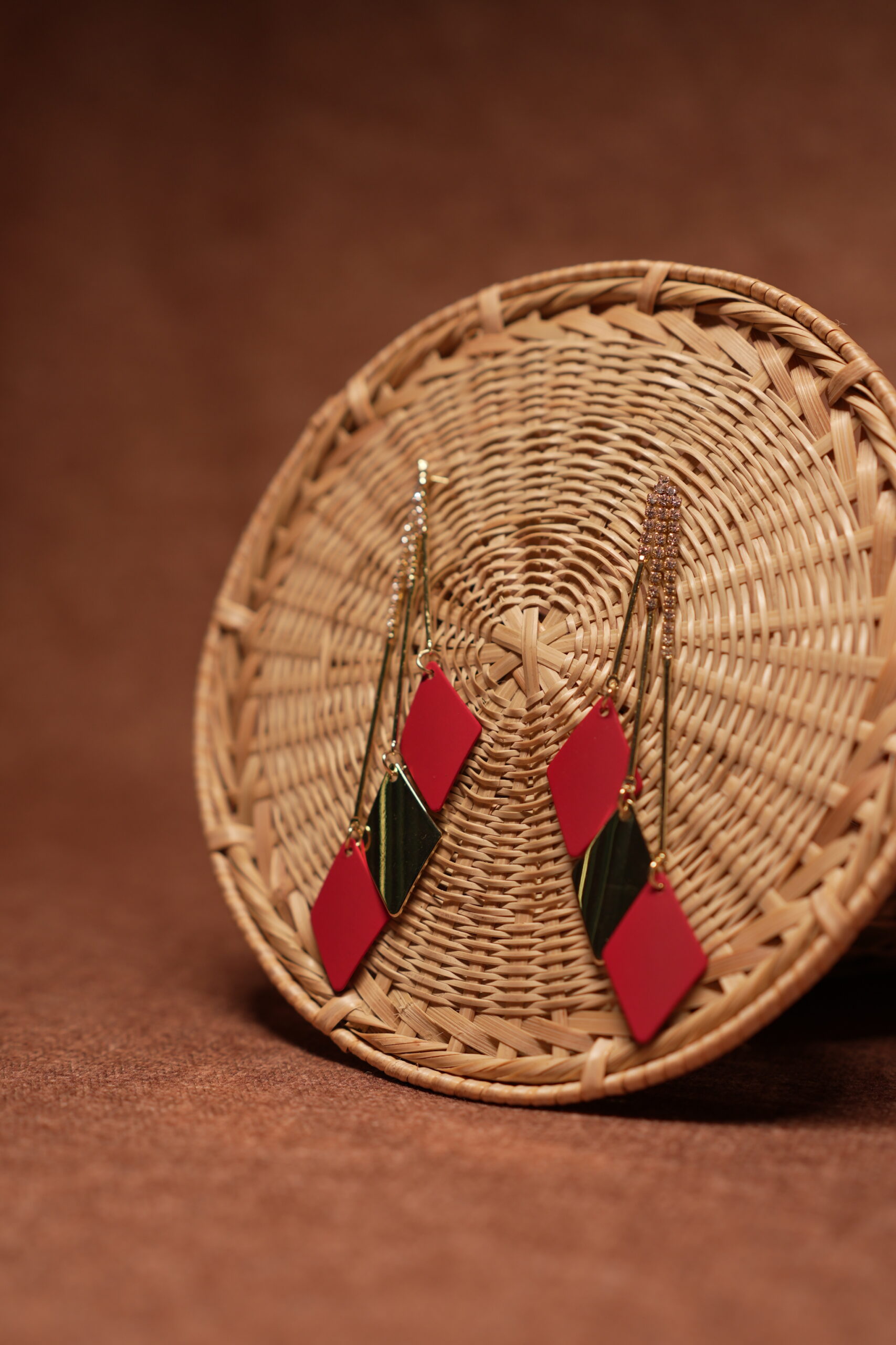 Red and green diamond shape layered drop earrings with long gold crystal chain tassel, displayed on a round wicker basket with warm brown background