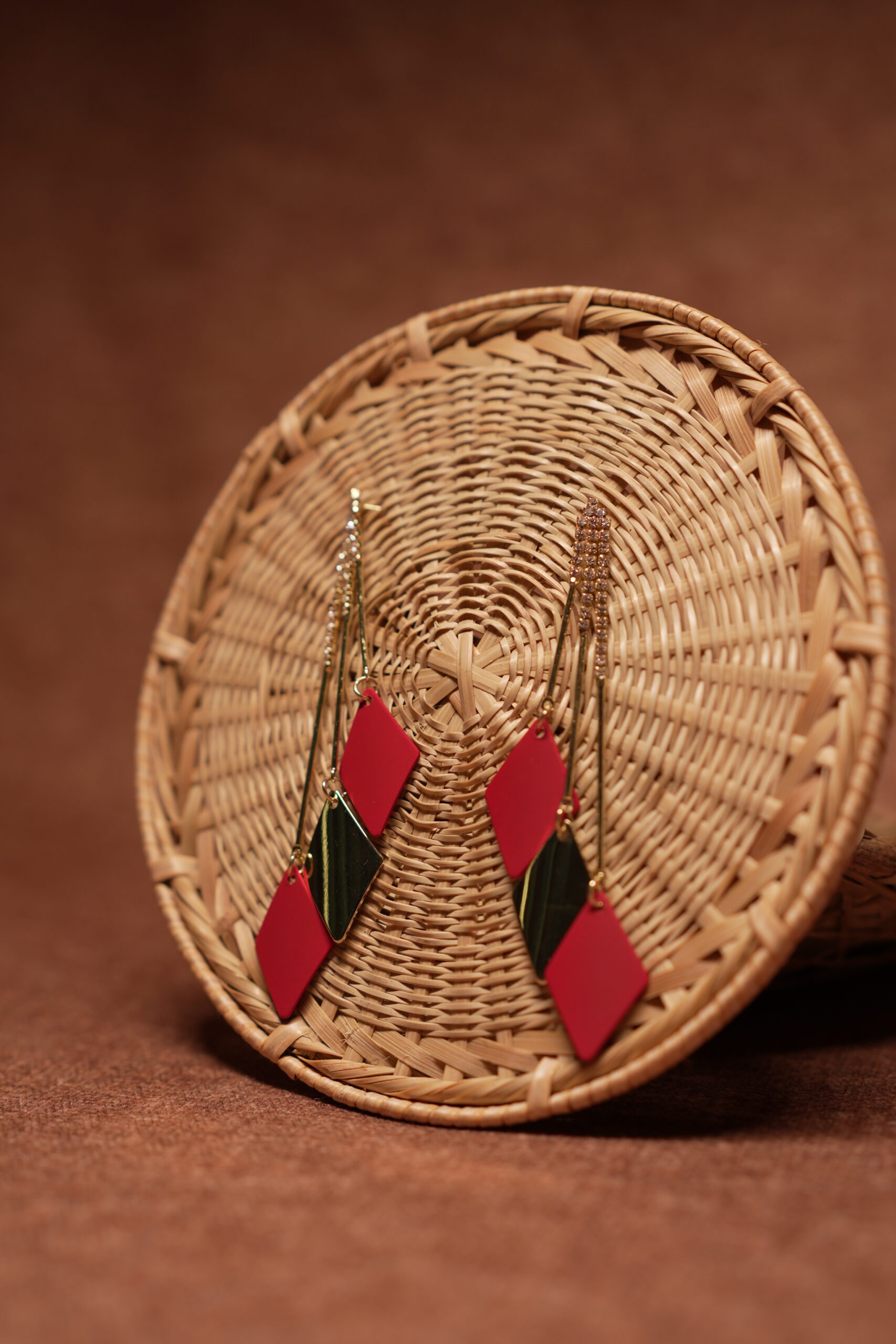 Red and green diamond shape layered drop earrings with long gold crystal chain tassel, displayed on a round wicker basket with warm brown background