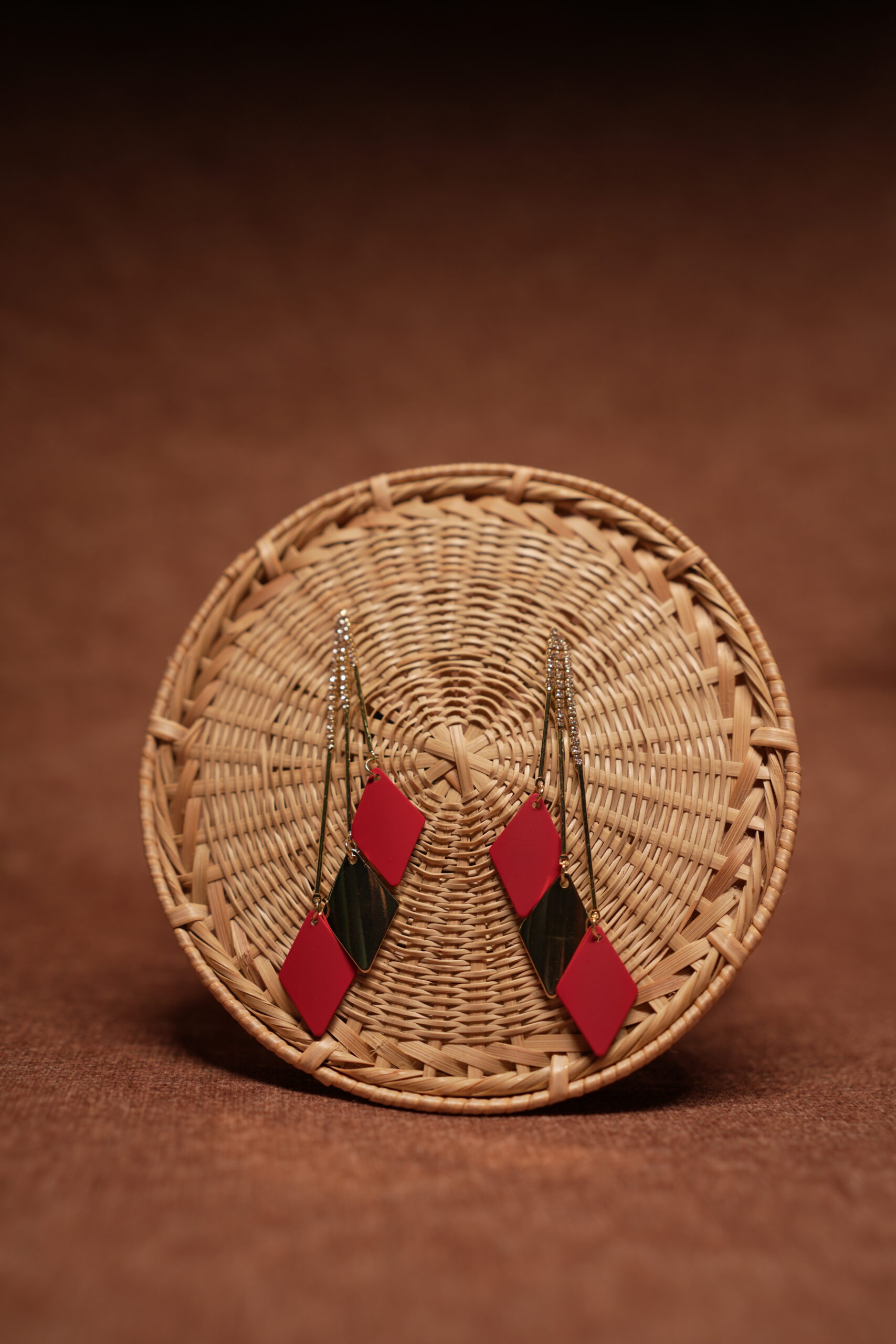 Red and green diamond shape layered drop earrings with long gold crystal chain tassel, displayed on a round wicker basket with warm brown background
