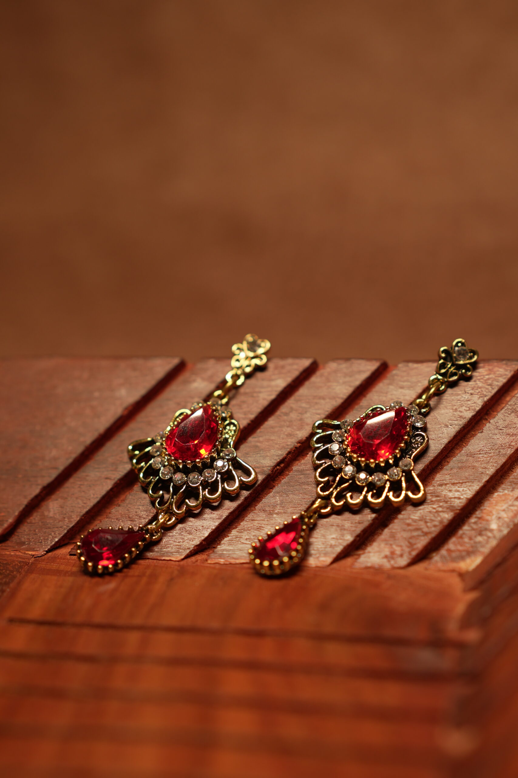 Antique gold floral filigree stud drop earrings with large ruby red teardrop crystal centre stone, white rhinestone accents and red teardrop pendant drop, displayed on wooden surface