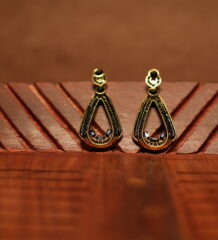 A pair of open teardrop drop earrings with a black crystal-encrusted frame, gold metal border, round black stone stud top, and baguette crystal accents at the base, displayed on a carved wooden surface.