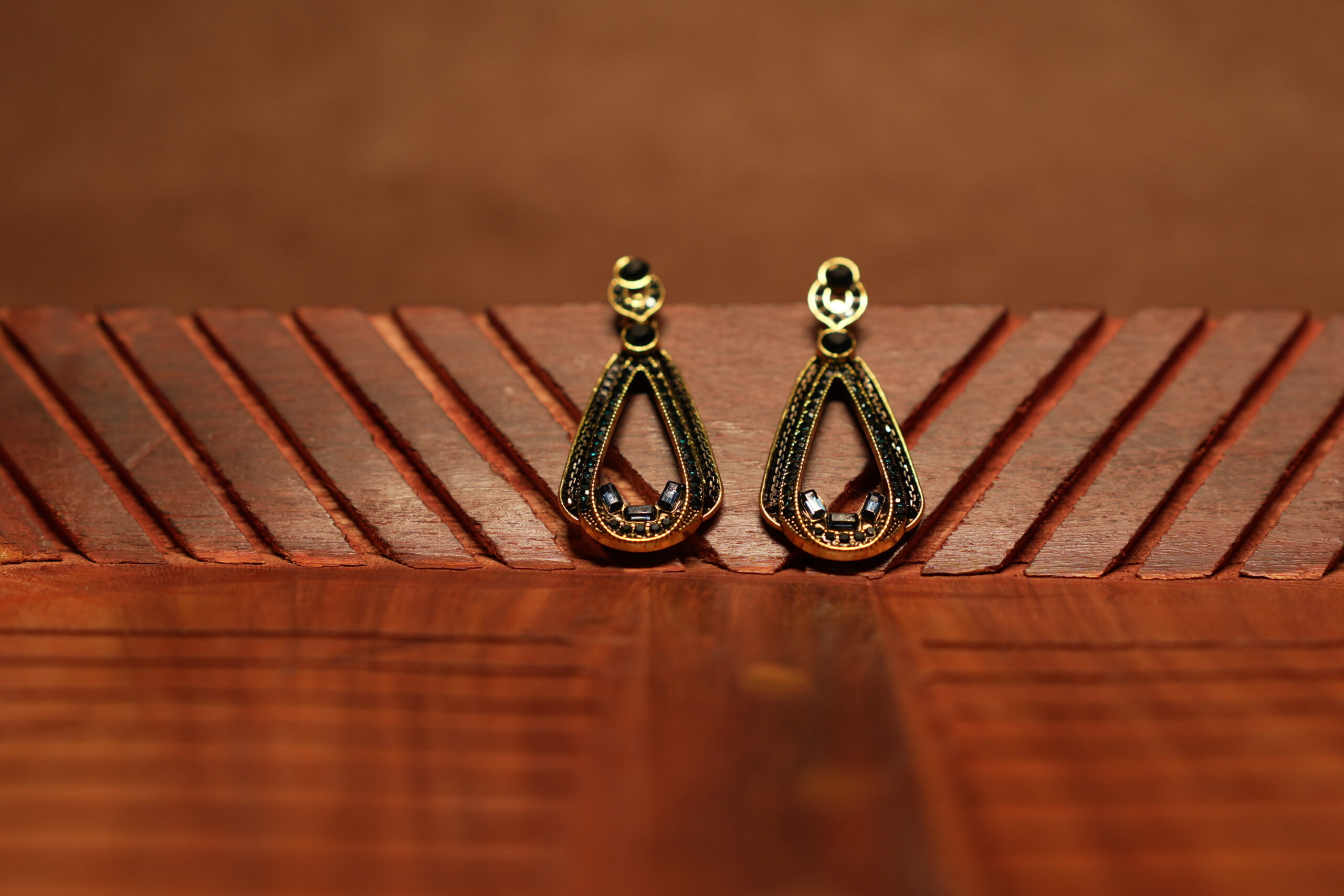 A pair of open teardrop drop earrings with a black crystal-encrusted frame, gold metal border, round black stone stud top, and baguette crystal accents at the base, displayed on a carved wooden surface.