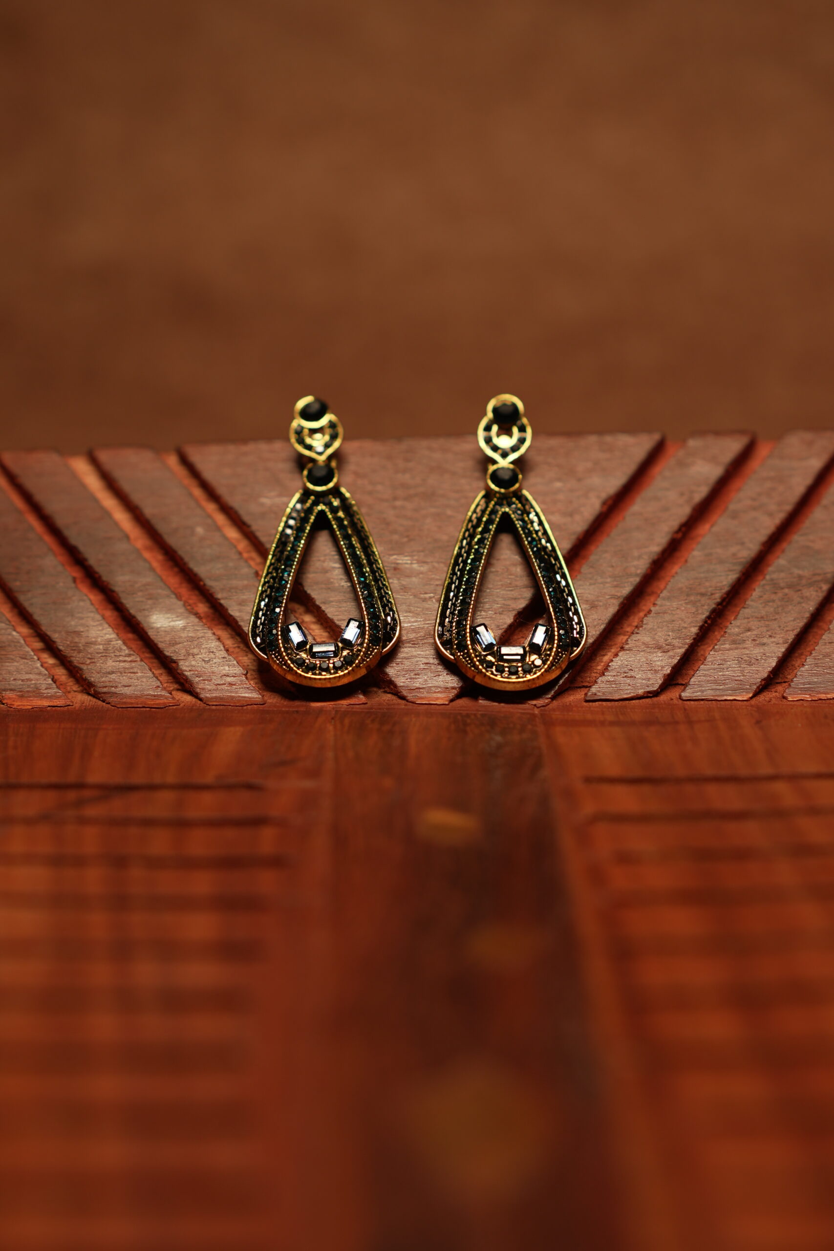 A pair of open teardrop drop earrings with a black crystal-encrusted frame, gold metal border, round black stone stud top, and baguette crystal accents at the base, displayed on a carved wooden surface.