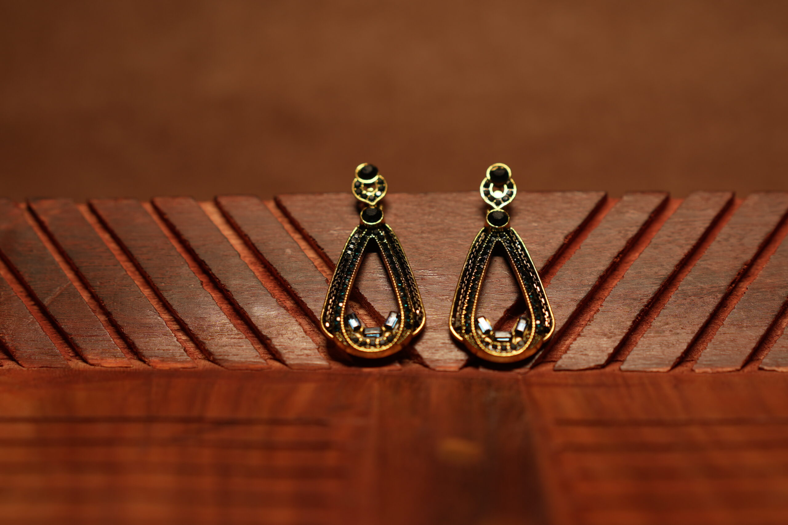 A pair of open teardrop drop earrings with a black crystal-encrusted frame, gold metal border, round black stone stud top, and baguette crystal accents at the base, displayed on a carved wooden surface.