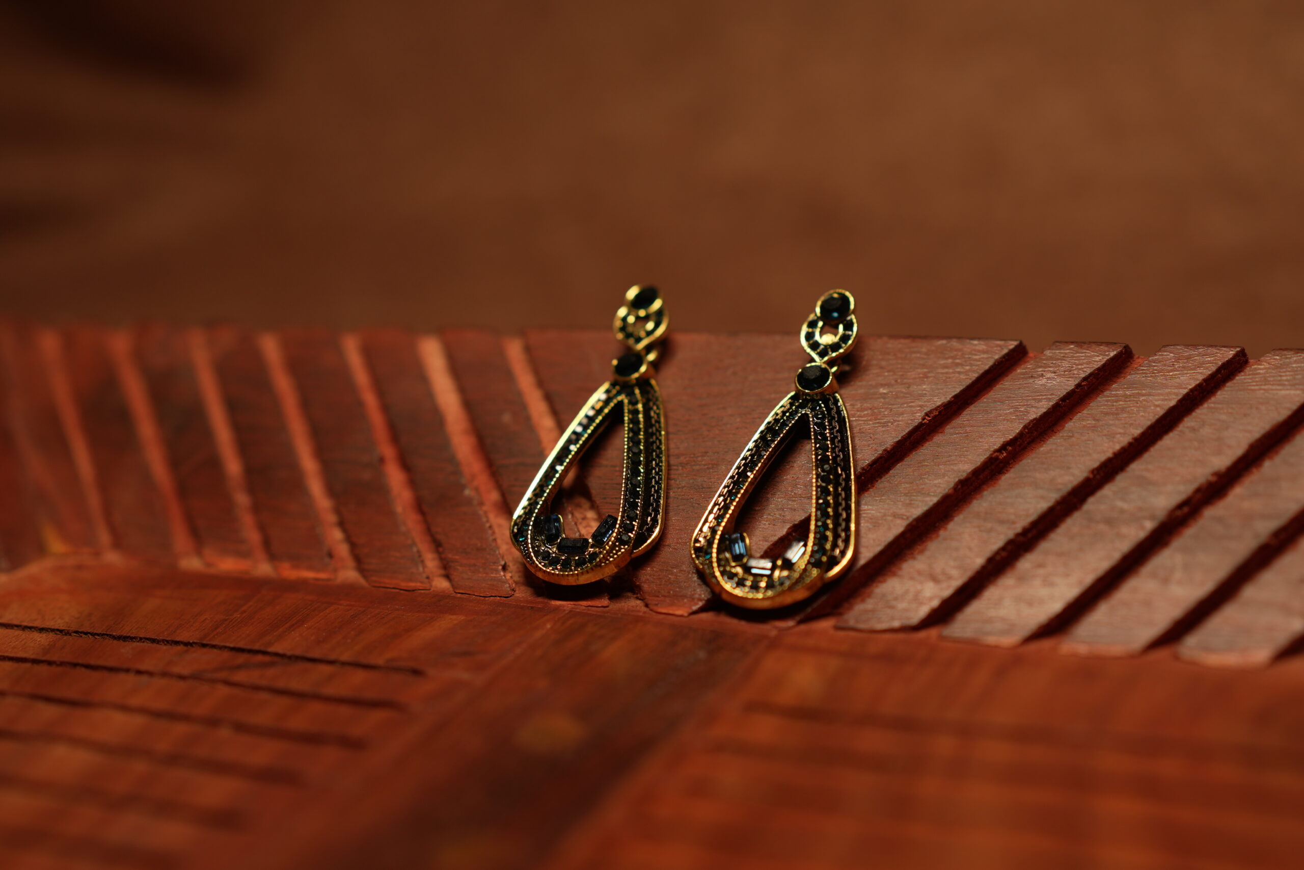 A pair of open teardrop drop earrings with a black crystal-encrusted frame, gold metal border, round black stone stud top, and baguette crystal accents at the base, displayed on a carved wooden surface.