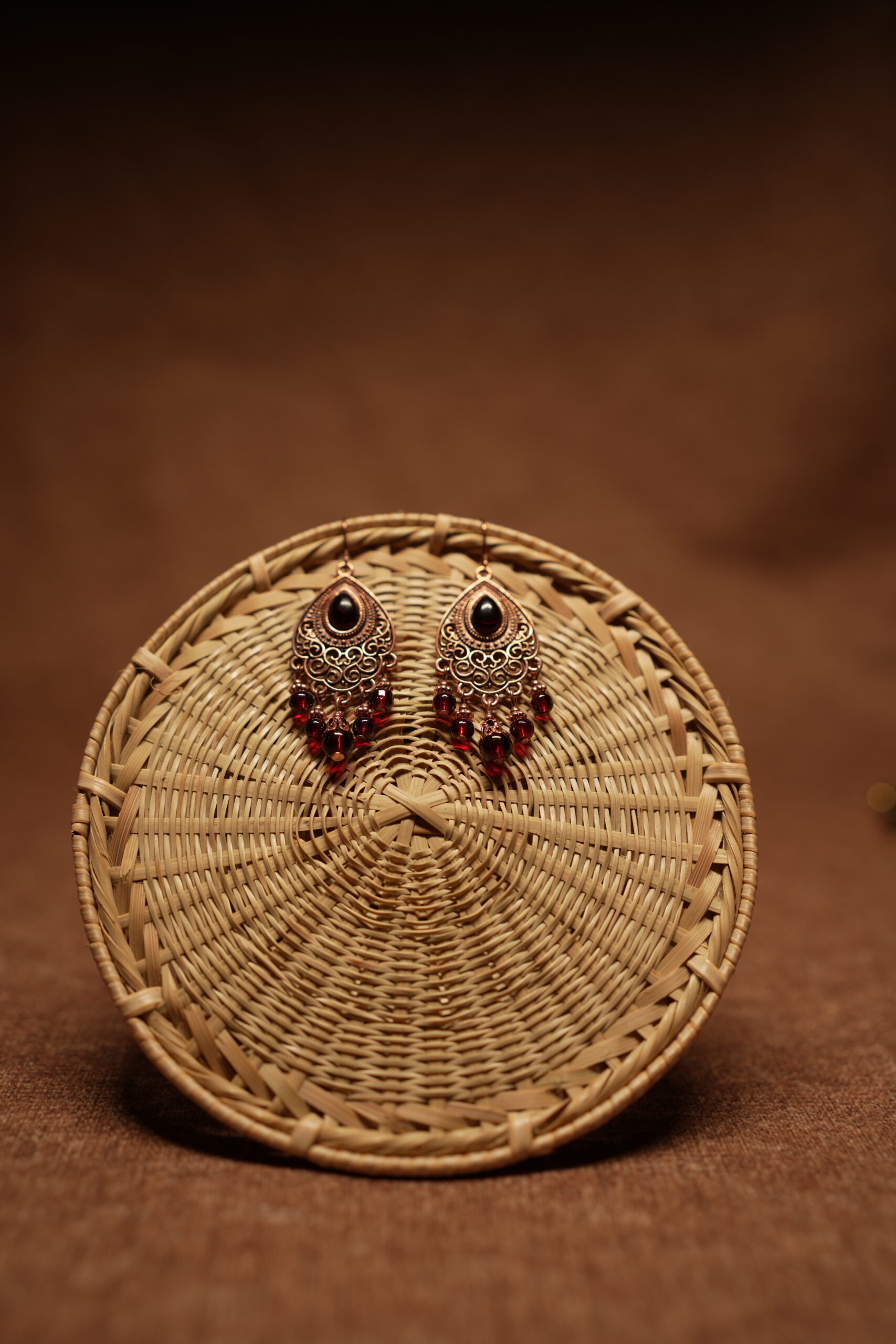 Antique copper filigree teardrop chandelier earrings with black onyx centre stone and red crystal bead drops, displayed on a round wicker basket with warm brown background