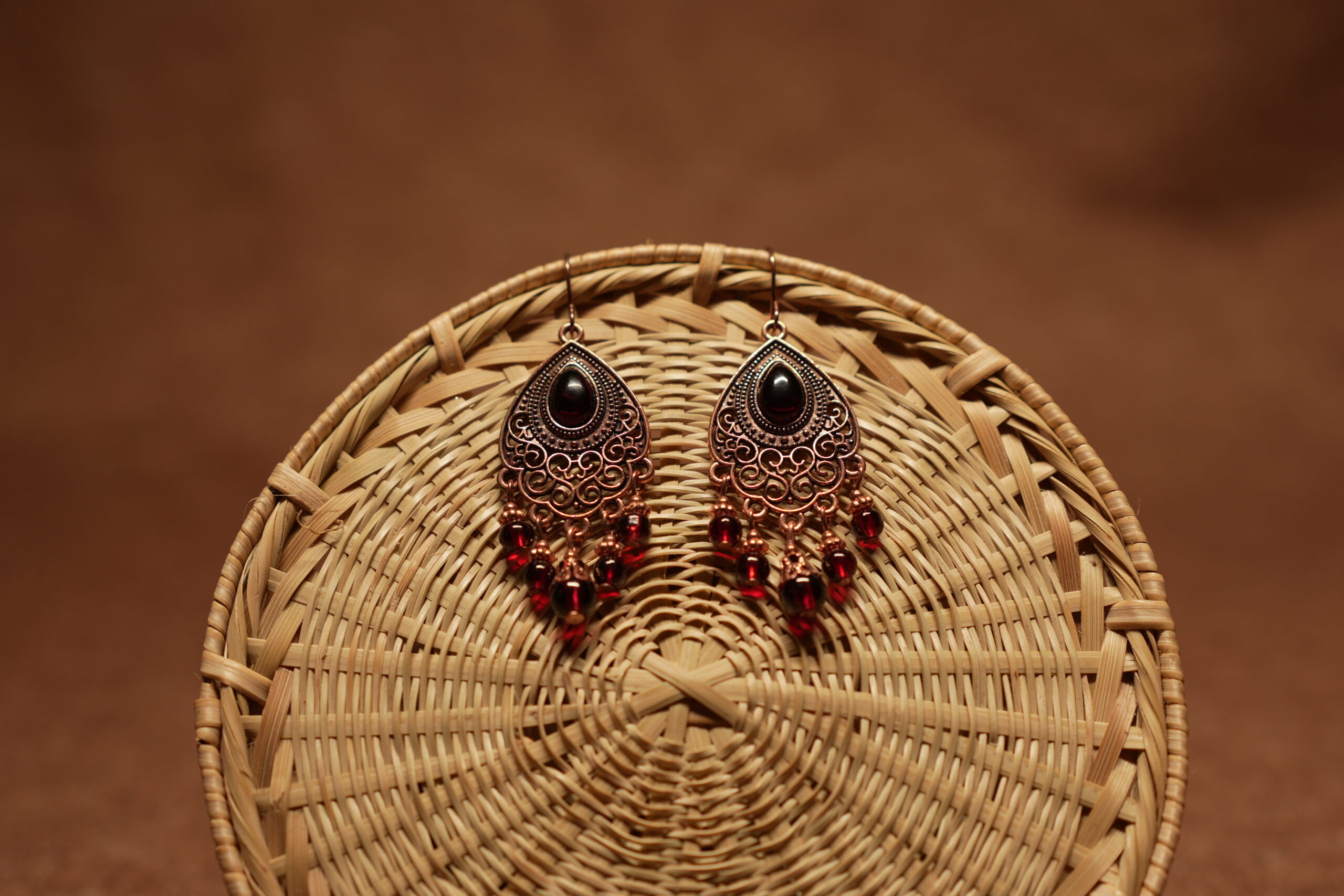 Antique copper filigree teardrop chandelier earrings with black onyx centre stone and red crystal bead drops, displayed on a round wicker basket with warm brown background