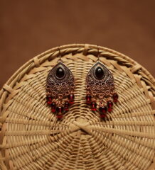 Antique copper filigree teardrop chandelier earrings with black onyx centre stone and red crystal bead drops, displayed on a round wicker basket with warm brown background
