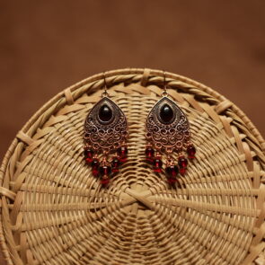 Antique copper filigree teardrop chandelier earrings with black onyx centre stone and red crystal bead drops, displayed on a round wicker basket with warm brown background