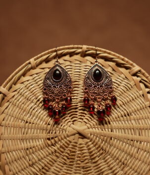 Antique copper filigree teardrop chandelier earrings with black onyx centre stone and red crystal bead drops, displayed on a round wicker basket with warm brown background