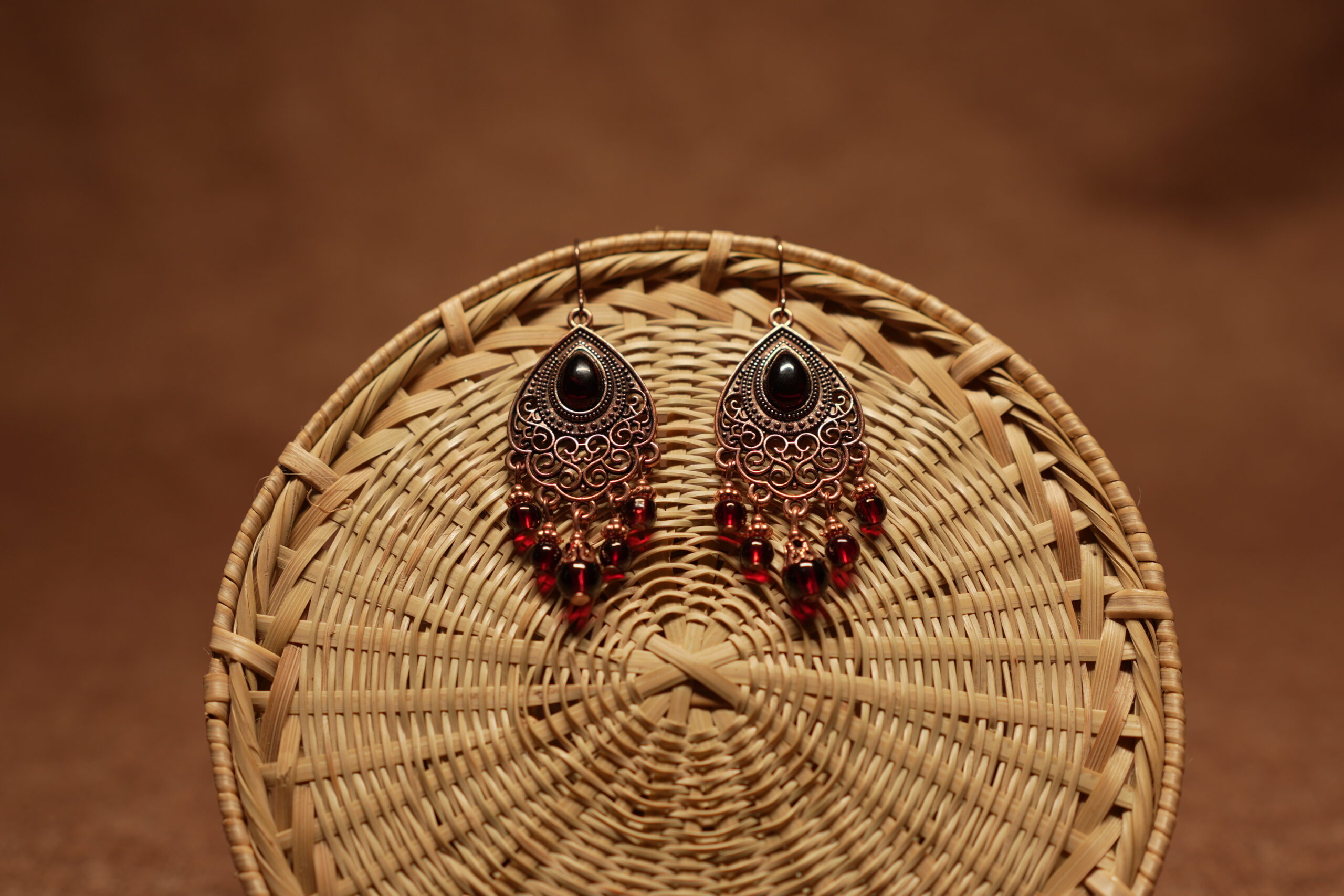 Antique copper filigree teardrop chandelier earrings with black onyx centre stone and red crystal bead drops, displayed on a round wicker basket with warm brown background