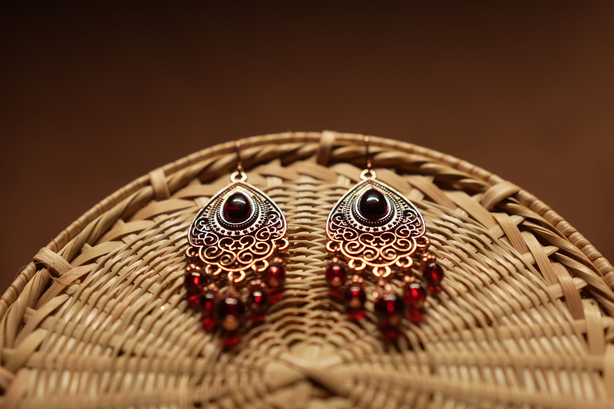 Antique copper filigree teardrop chandelier earrings with black onyx centre stone and red crystal bead drops, displayed on a round wicker basket with warm brown background