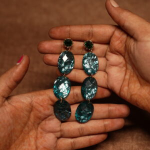 Long triple drop earrings featuring three graduated faceted oval teal stones in an ombre from light aqua to deep teal, topped with a dark green crystal stud in a gold claw setting, held in a woman's open palms.