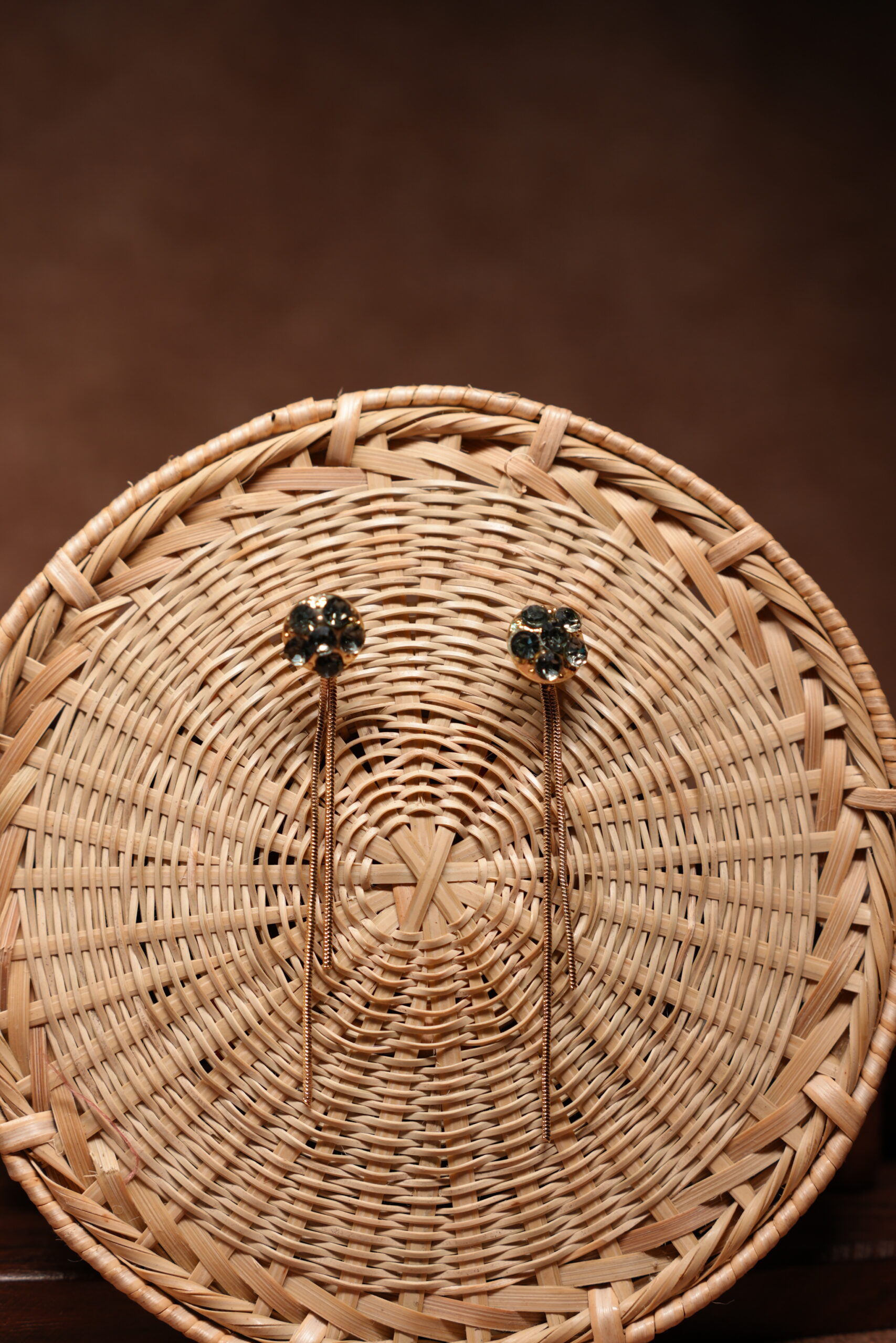 Gold-tone floral dark crystal cluster stud earrings with long multi-strand chain tassel drops, displayed on a round woven rattan basket against a brown background