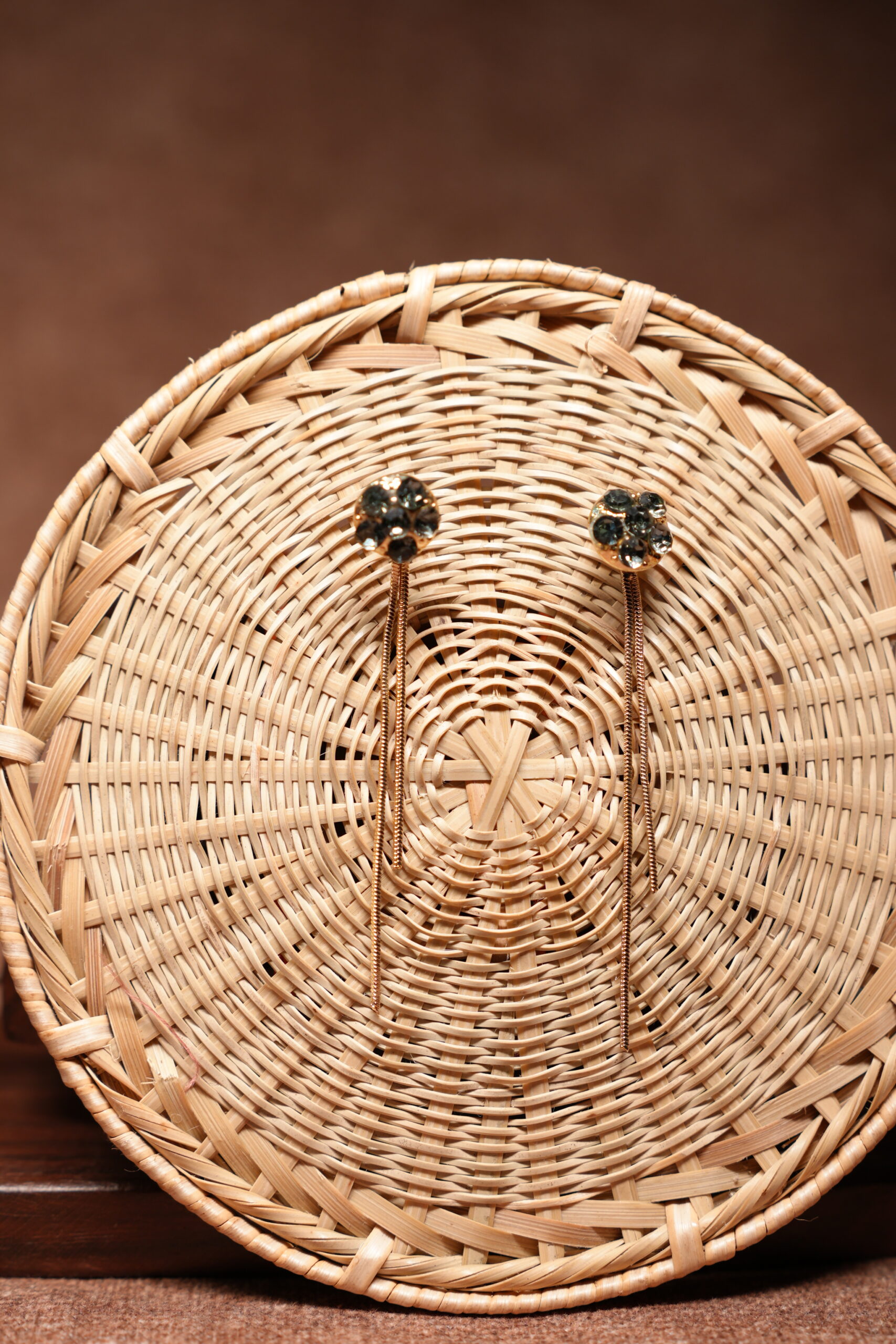 Gold-tone floral dark crystal cluster stud earrings with long multi-strand chain tassel drops, displayed on a round woven rattan basket against a brown background