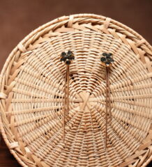 Gold-tone floral dark crystal cluster stud earrings with long multi-strand chain tassel drops, displayed on a round woven rattan basket against a brown background