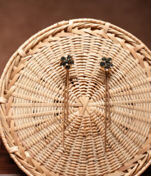 Gold-tone floral dark crystal cluster stud earrings with long multi-strand chain tassel drops, displayed on a round woven rattan basket against a brown background