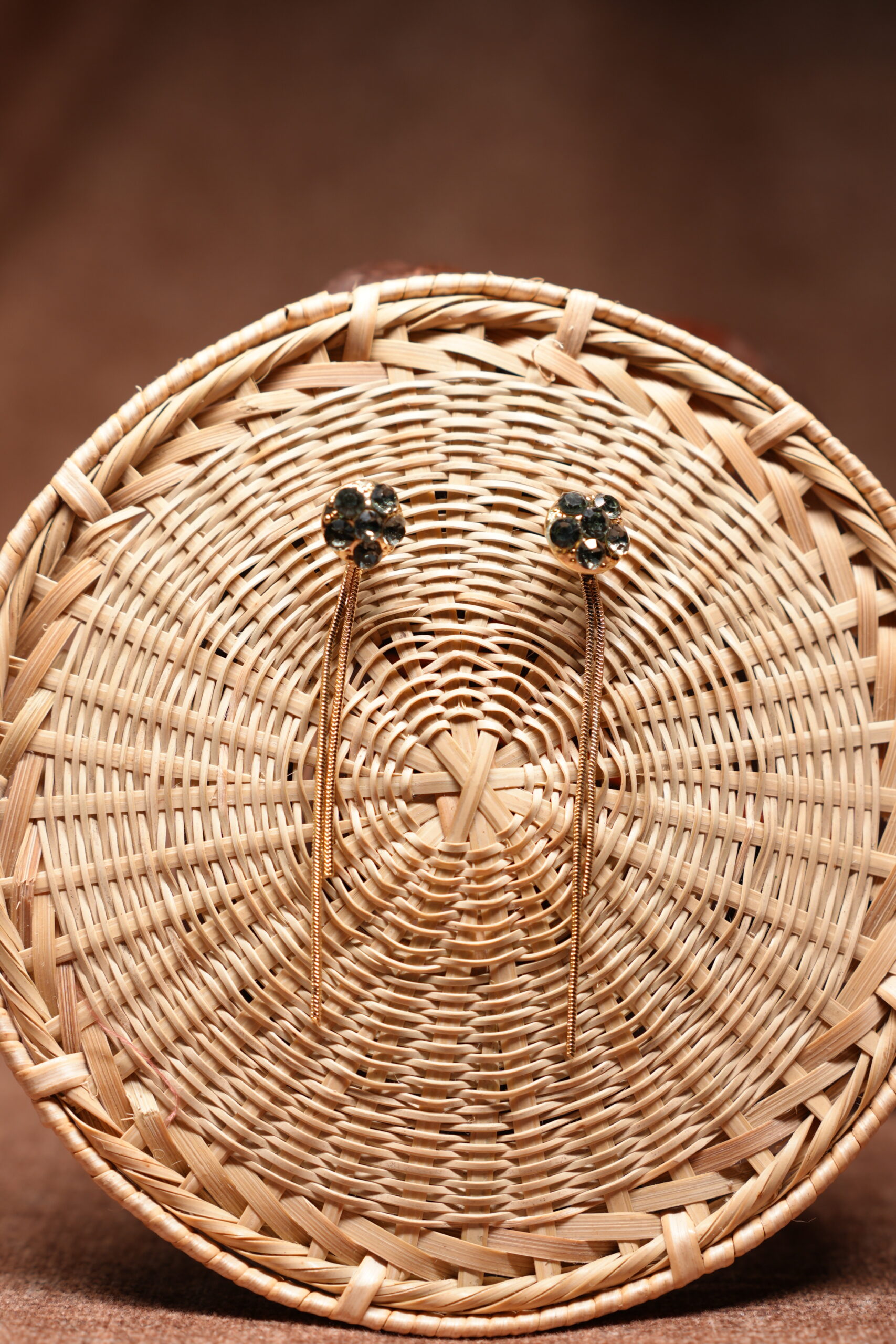 Gold-tone floral dark crystal cluster stud earrings with long multi-strand chain tassel drops, displayed on a round woven rattan basket against a brown background