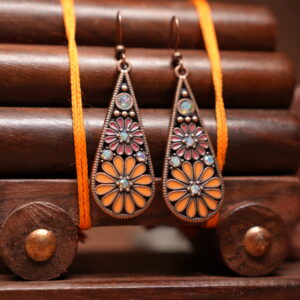Antique copper-toned teardrop dangle earrings with orange and pink enamel floral motif and opal-effect stone accents, hung on a wooden jewellery stand with a carved elephant figurine
