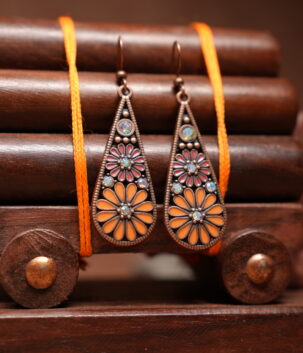 Antique copper-toned teardrop dangle earrings with orange and pink enamel floral motif and opal-effect stone accents, hung on a wooden jewellery stand with a carved elephant figurine