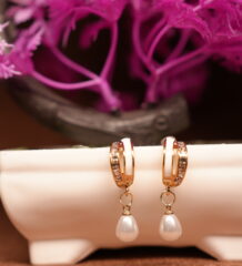Gold-toned crystal huggie hoop earrings with white teardrop pearl charm, displayed on a white ceramic stand with pink flowers in the background