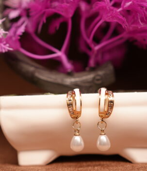 Gold-toned crystal huggie hoop earrings with white teardrop pearl charm, displayed on a white ceramic stand with pink flowers in the background