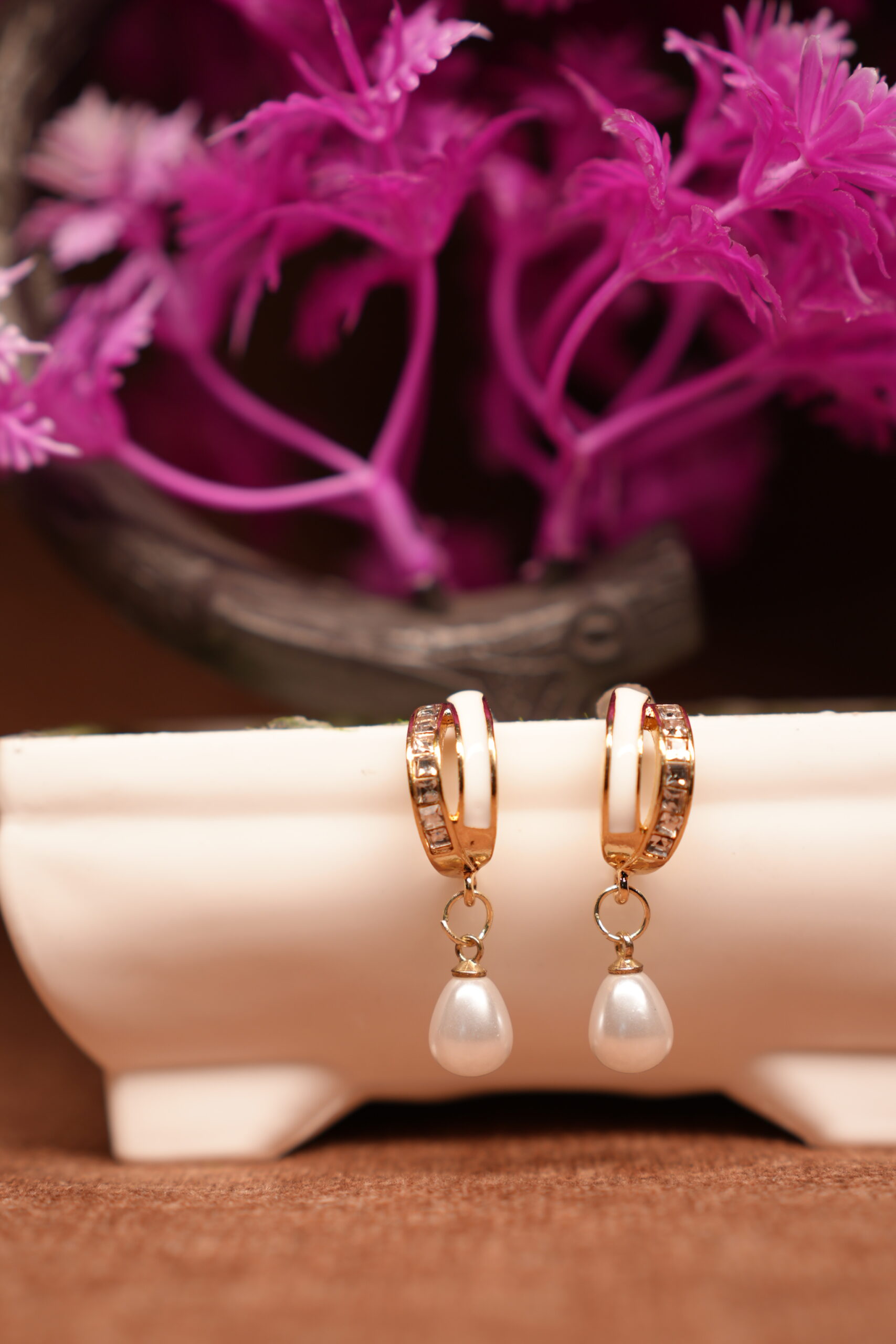 Gold-toned crystal huggie hoop earrings with white teardrop pearl charm, displayed on a white ceramic stand with pink flowers in the background