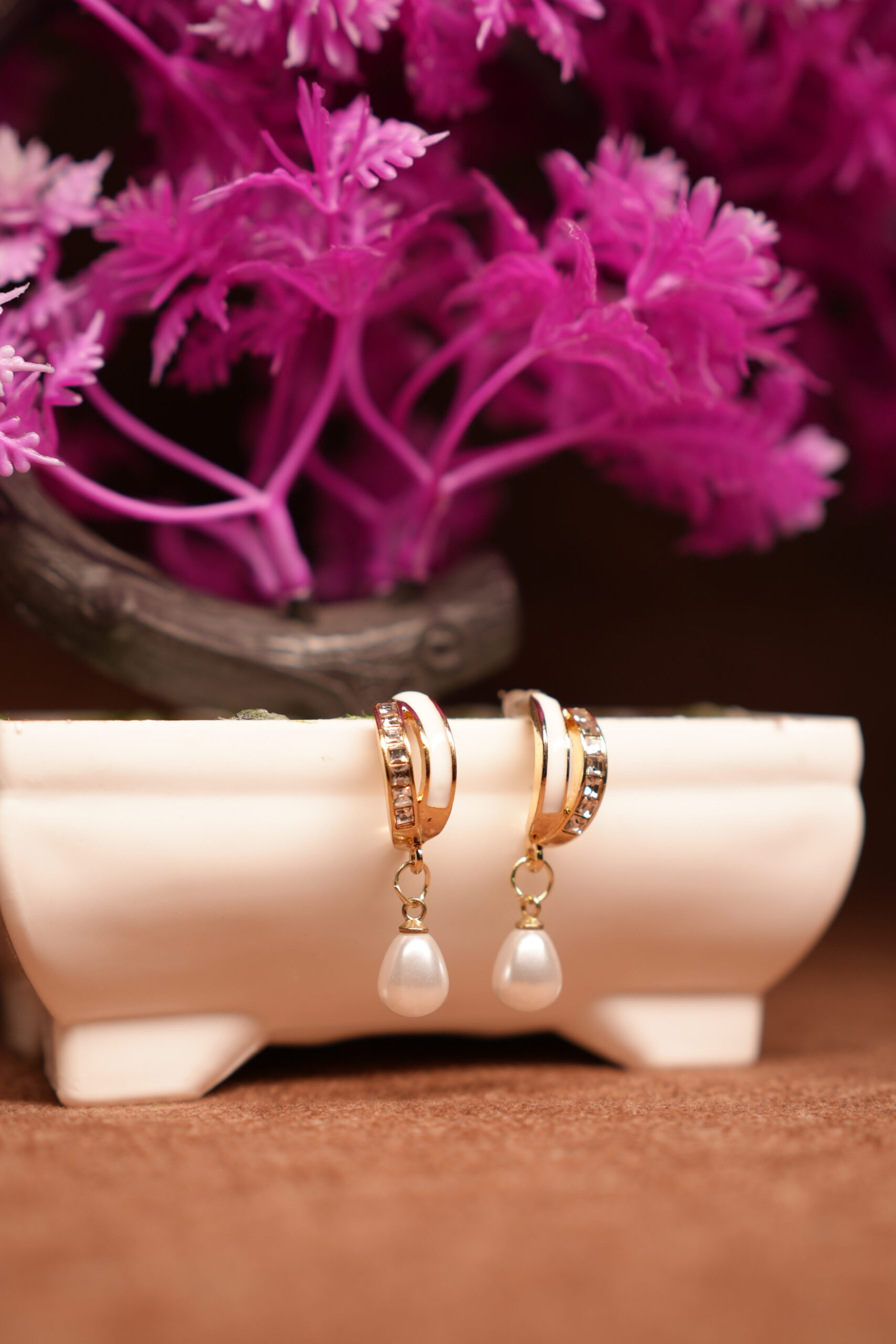 Gold-toned crystal huggie hoop earrings with white teardrop pearl charm, displayed on a white ceramic stand with pink flowers in the background