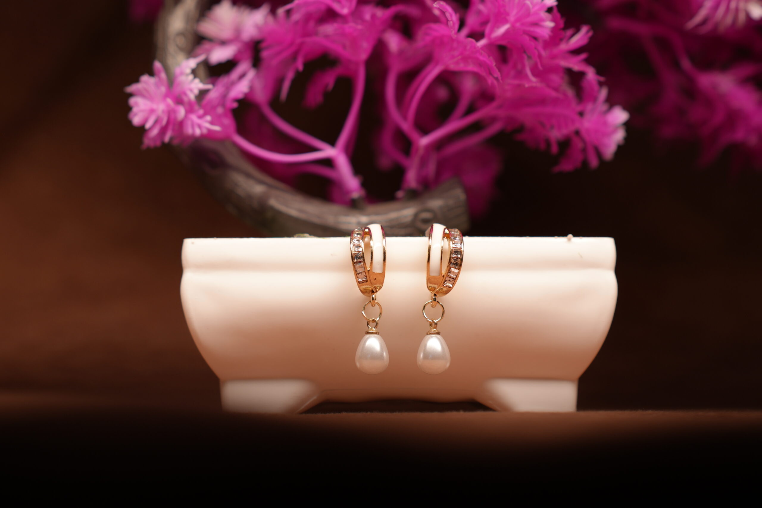 Gold-toned crystal huggie hoop earrings with white teardrop pearl charm, displayed on a white ceramic stand with pink flowers in the background