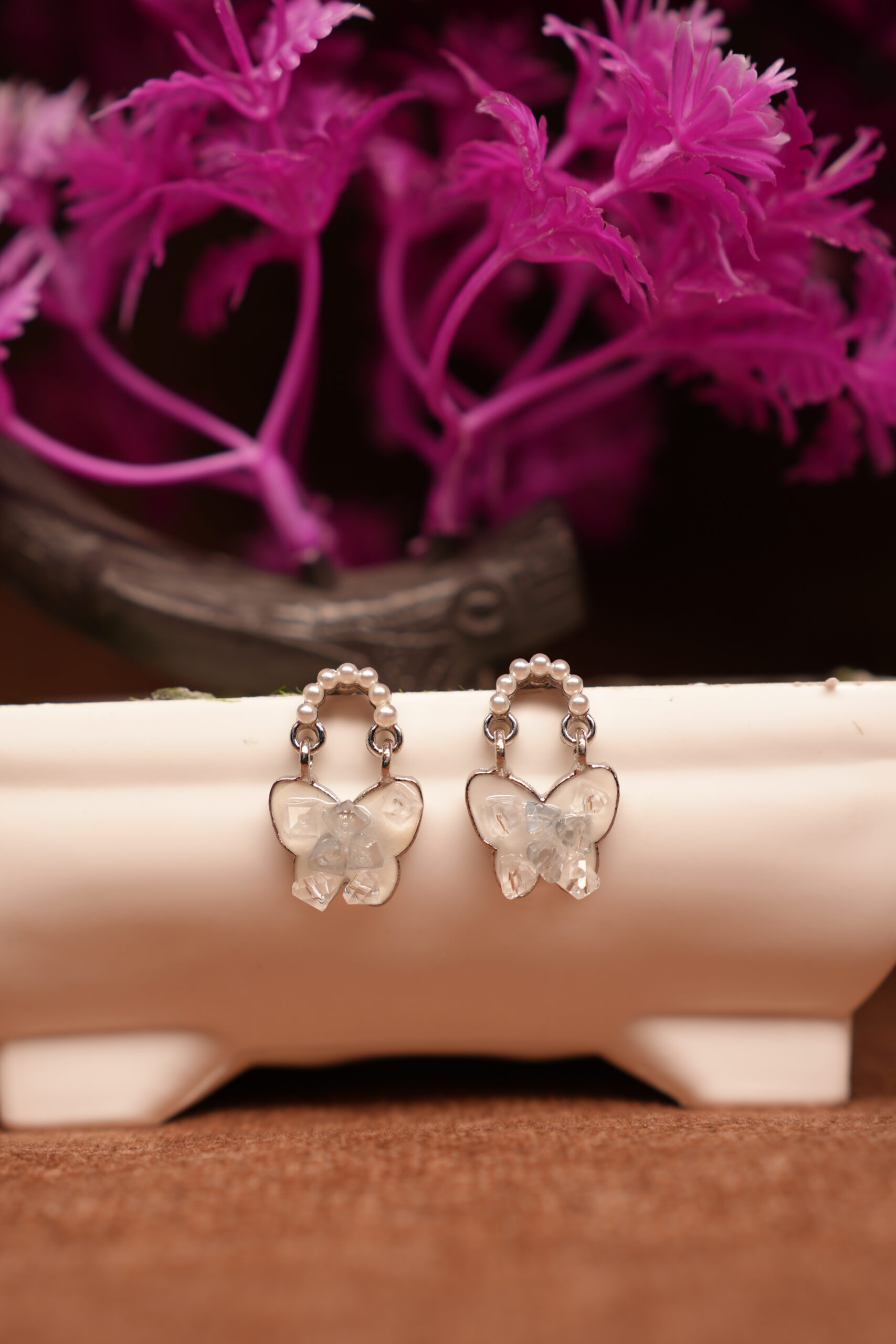 Silver pearl hoop earrings with clear crystal butterfly pendant drop displayed on white ceramic stand with pink flowers – D2Dazzle fashion jewellery India