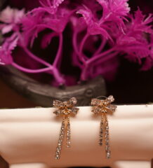 Gold-tone rhinestone bow stud earrings with triple crystal tassel chains displayed on white ceramic stand with pink flowers – D2Dazzle fashion jewellery India