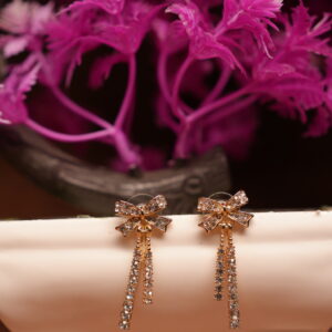 Gold-tone rhinestone bow stud earrings with triple crystal tassel chains displayed on white ceramic stand with pink flowers – D2Dazzle fashion jewellery India