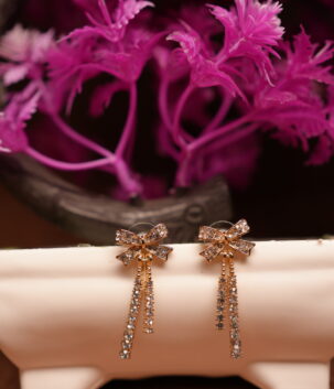 Gold-tone rhinestone bow stud earrings with triple crystal tassel chains displayed on white ceramic stand with pink flowers – D2Dazzle fashion jewellery India