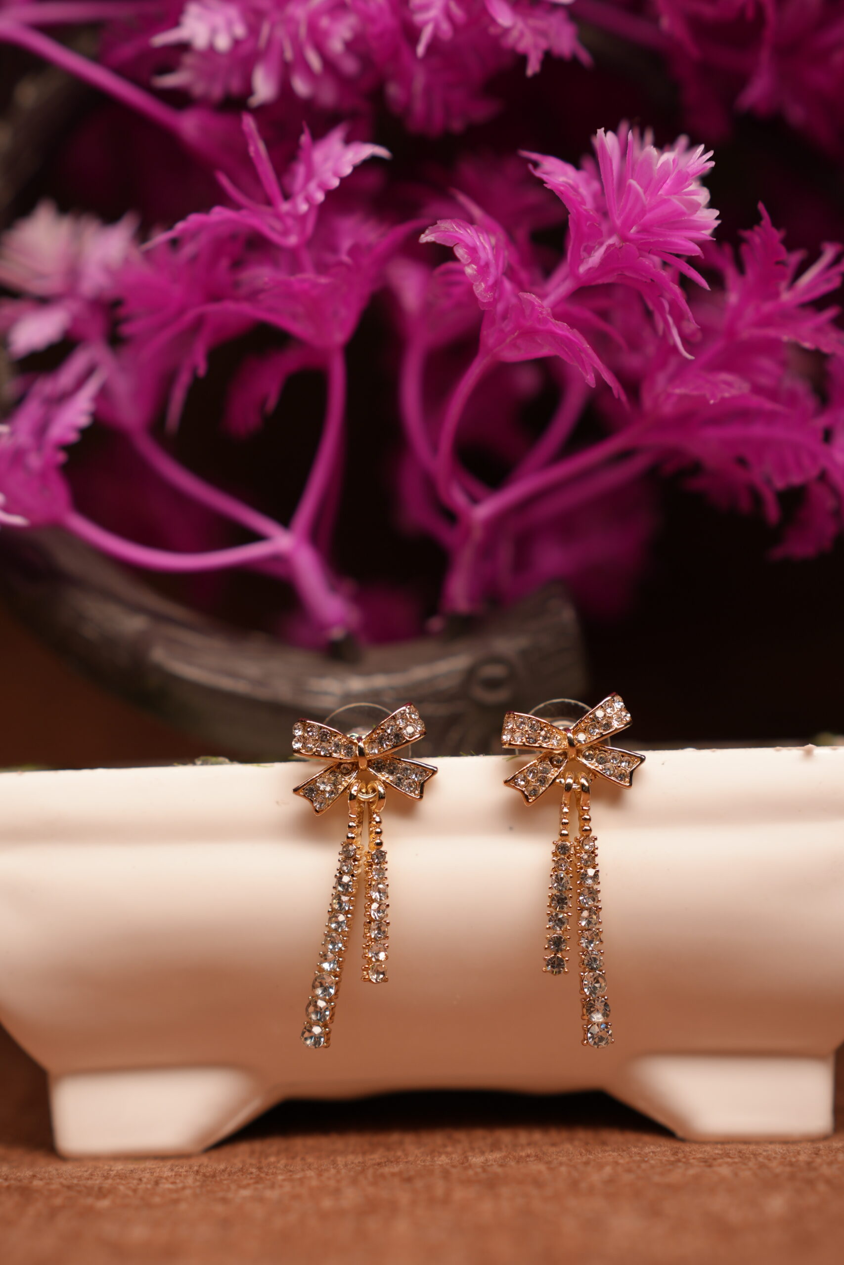 Gold-tone rhinestone bow stud earrings with triple crystal tassel chains displayed on white ceramic stand with pink flowers – D2Dazzle fashion jewellery India