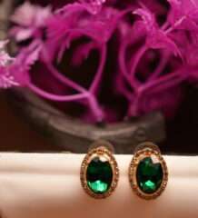 Emerald green oval halo stud earrings with gold-tone rhinestone crystal border displayed on white ceramic stand with pink flowers – D2Dazzle fashion jewellery India