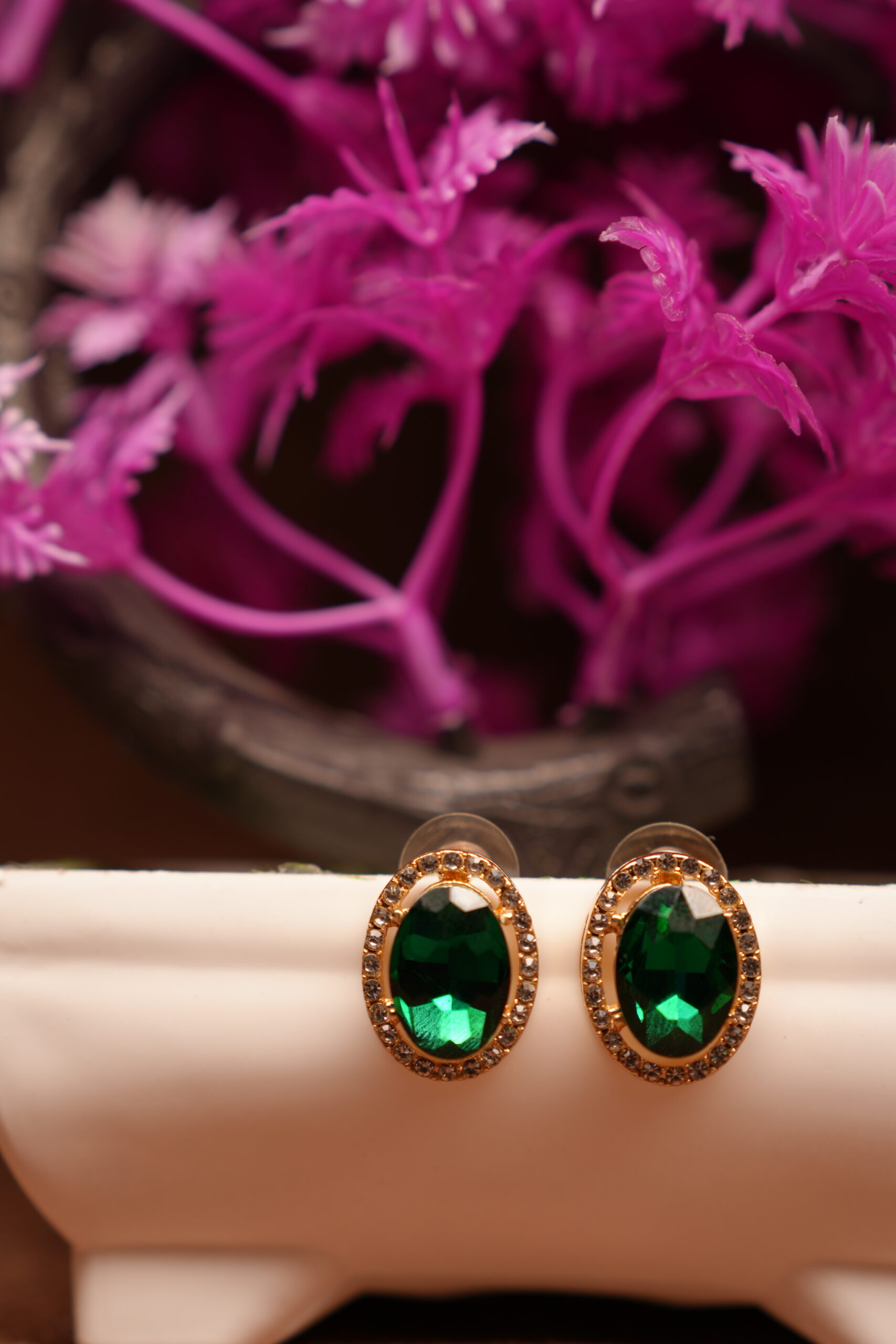 Emerald green oval halo stud earrings with gold-tone rhinestone crystal border displayed on white ceramic stand with pink flowers – D2Dazzle fashion jewellery India