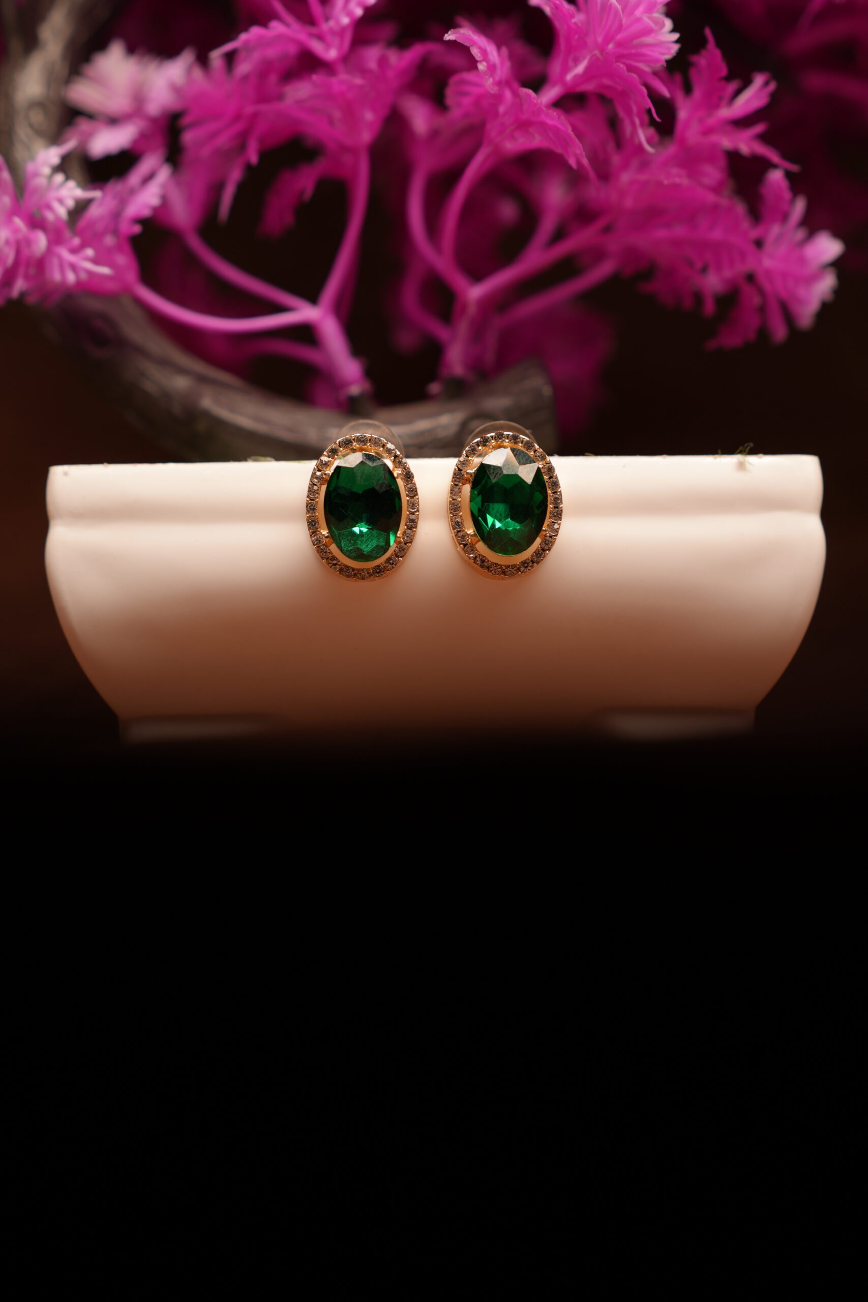 Emerald green oval halo stud earrings with gold-tone rhinestone crystal border displayed on white ceramic stand with pink flowers – D2Dazzle fashion jewellery India