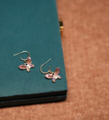 A pair of gold-tone butterfly drop earrings featuring pink marquise crystal wings and an iridescent oval centre stone, placed on a teal velvet jewellery box.