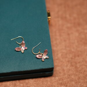 A pair of gold-tone butterfly drop earrings featuring pink marquise crystal wings and an iridescent oval centre stone, placed on a teal velvet jewellery box.
