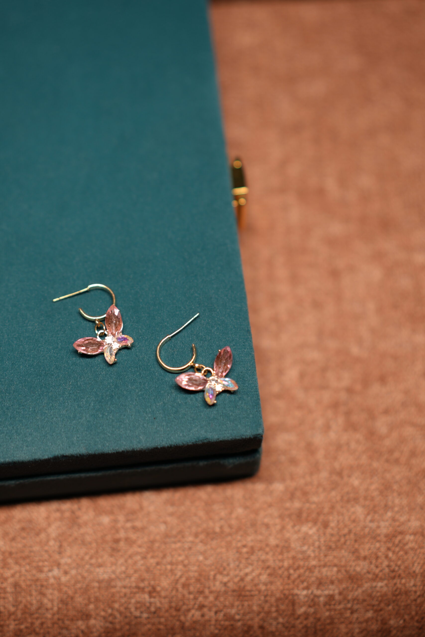 A pair of gold-tone butterfly drop earrings featuring pink marquise crystal wings and an iridescent oval centre stone, placed on a teal velvet jewellery box.