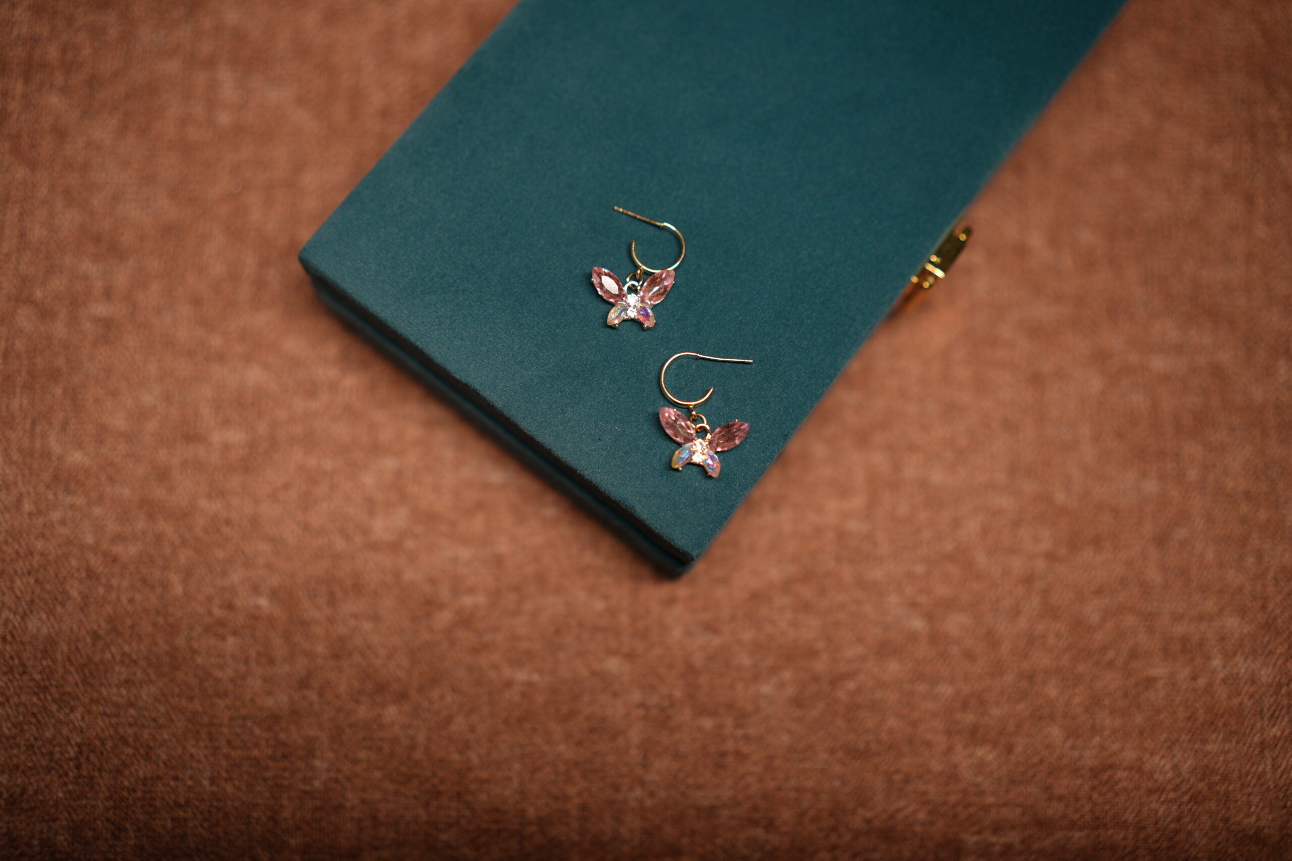 A pair of gold-tone butterfly drop earrings featuring pink marquise crystal wings and an iridescent oval centre stone, placed on a teal velvet jewellery box.