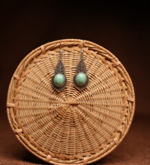 A pair of antique-silver boho leaf drop earrings with oval turquoise cabochon stones, displayed against a round woven rattan disc on a warm brown background.