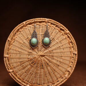 A pair of antique-silver boho leaf drop earrings with oval turquoise cabochon stones, displayed against a round woven rattan disc on a warm brown background.