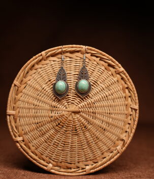 A pair of antique-silver boho leaf drop earrings with oval turquoise cabochon stones, displayed against a round woven rattan disc on a warm brown background.