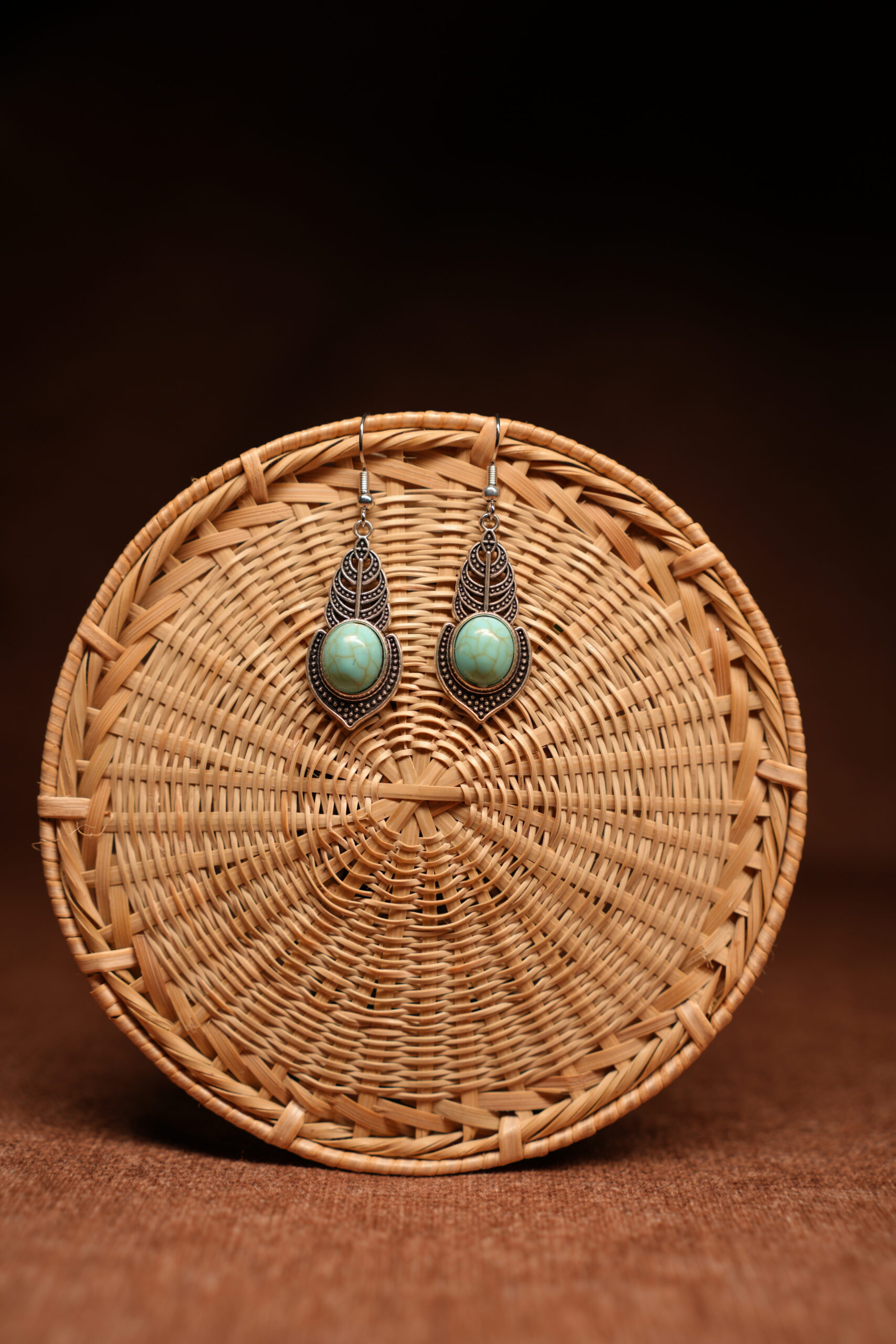 A pair of antique-silver boho leaf drop earrings with oval turquoise cabochon stones, displayed against a round woven rattan disc on a warm brown background.