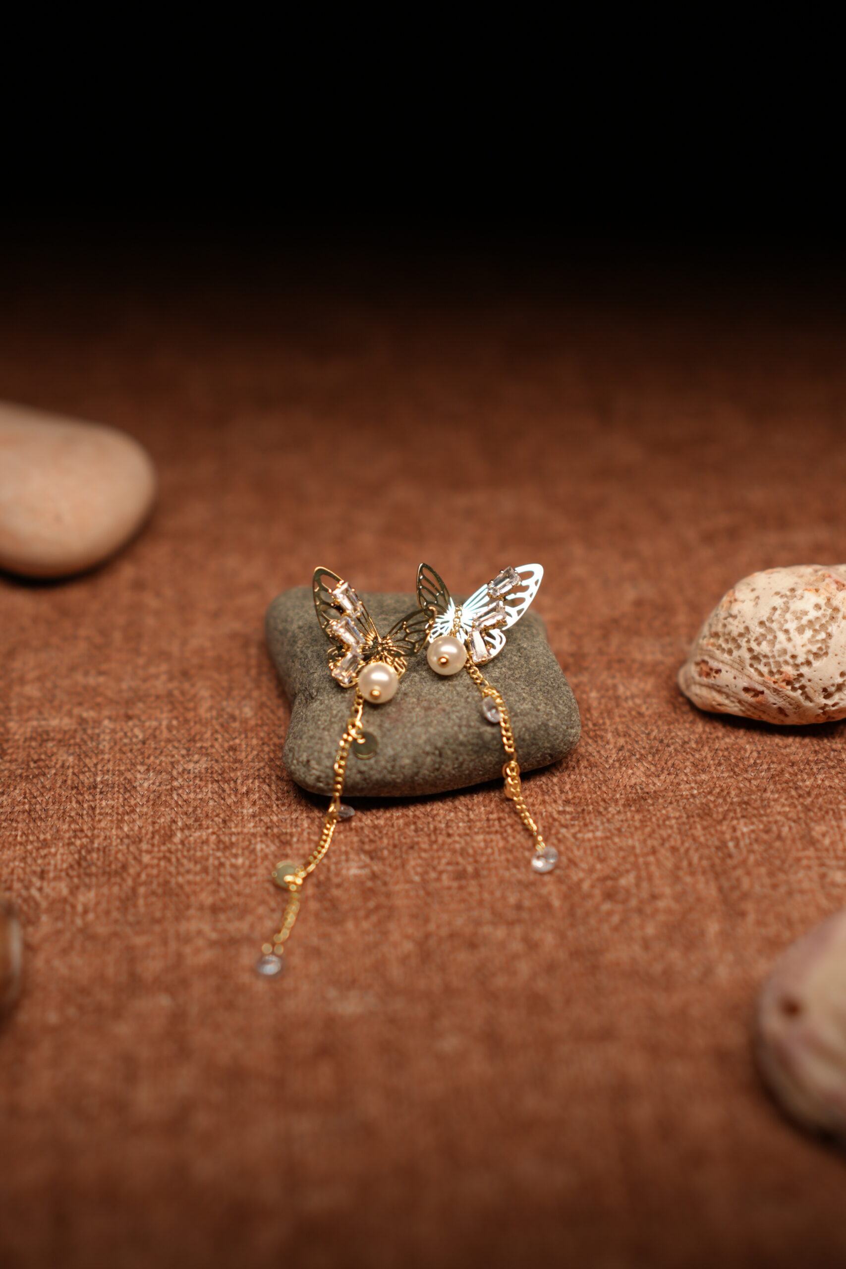 A pair of gold-tone filigree butterfly earrings with baguette crystals, faux pearl centres, and three delicate crystal-tipped chain tassels, resting on a smooth grey pebble against a warm brown fabric background.