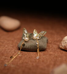 A pair of gold-tone filigree butterfly earrings with baguette crystals, faux pearl centres, and three delicate crystal-tipped chain tassels, resting on a smooth grey pebble against a warm brown fabric background.