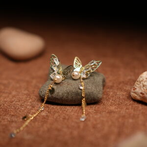 A pair of gold-tone filigree butterfly earrings with baguette crystals, faux pearl centres, and three delicate crystal-tipped chain tassels, resting on a smooth grey pebble against a warm brown fabric background.