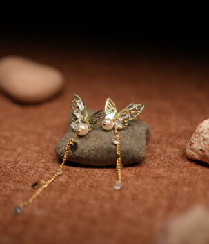 A pair of gold-tone filigree butterfly earrings with baguette crystals, faux pearl centres, and three delicate crystal-tipped chain tassels, resting on a smooth grey pebble against a warm brown fabric background.