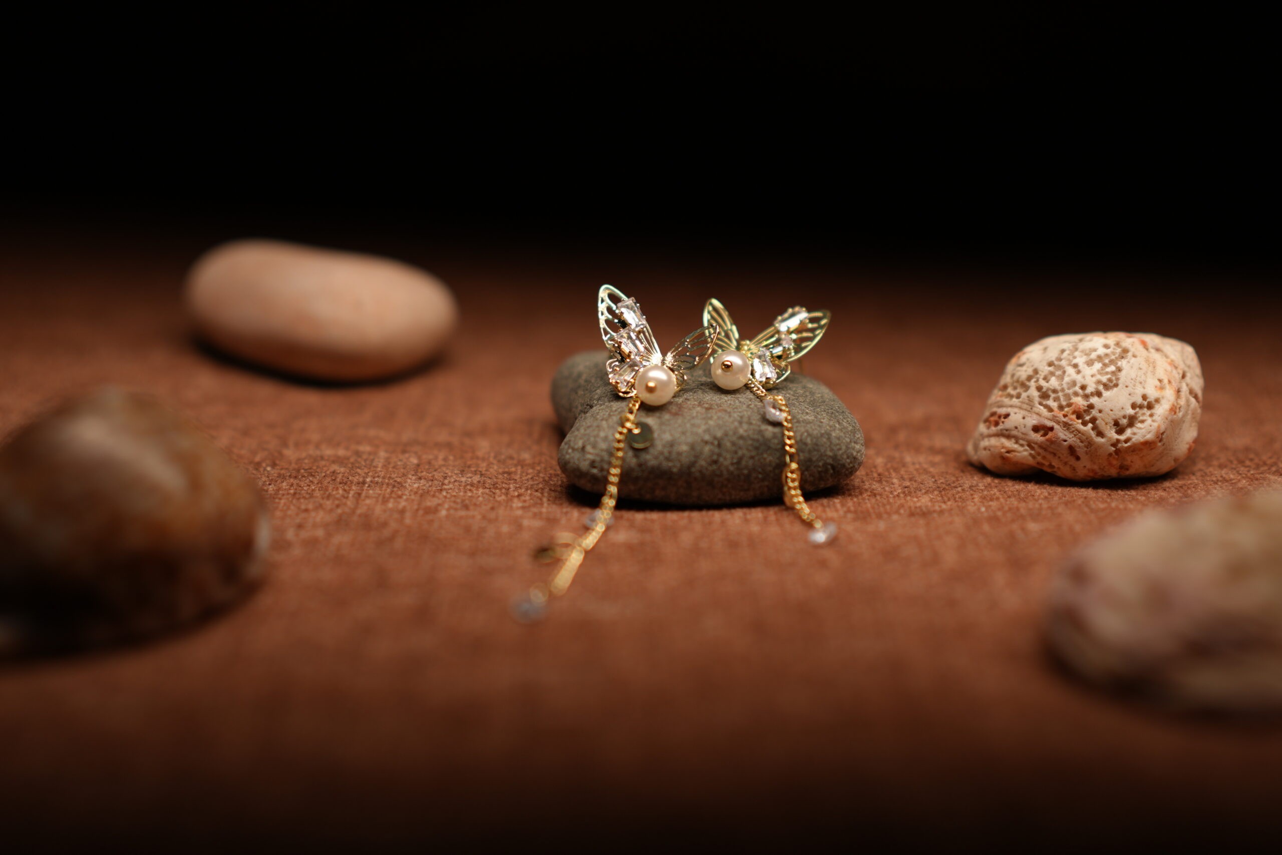 A pair of gold-tone filigree butterfly earrings with baguette crystals, faux pearl centres, and three delicate crystal-tipped chain tassels, resting on a smooth grey pebble against a warm brown fabric background.